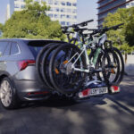 Naložená elektrokola na nosiči tažného zařízení u šedého auta Škoda před cestou.