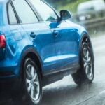 Akční pohled na zadní část jedoucího modrého SUV, jak jeho pneumatiky za deště odhazují vodu, což ilustruje okamžik před ztrátou přilnavosti a vznikem aquaplaningu.